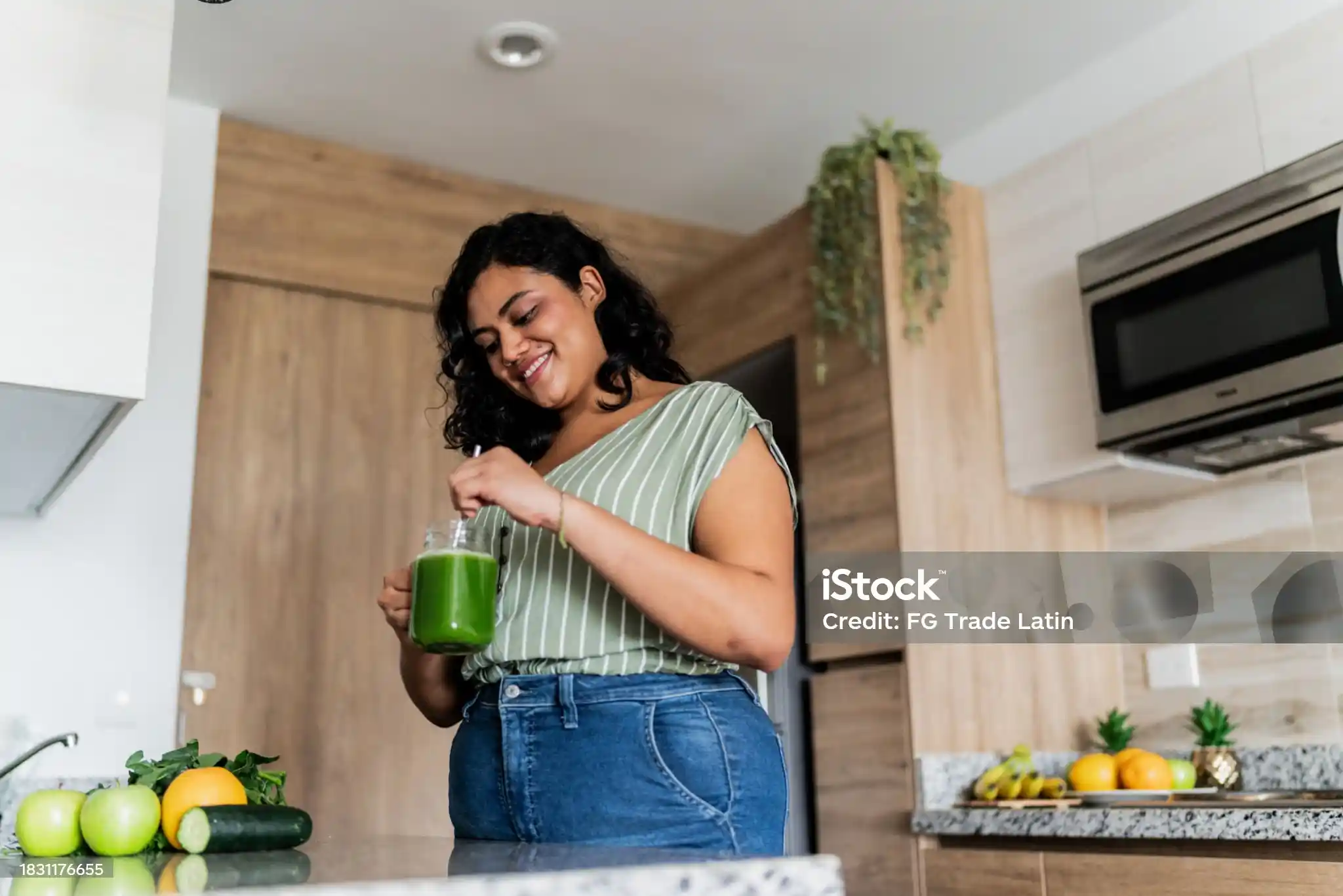Mujer sonriente preparando un batido verde en su cocina, rodeada de fruta fresca